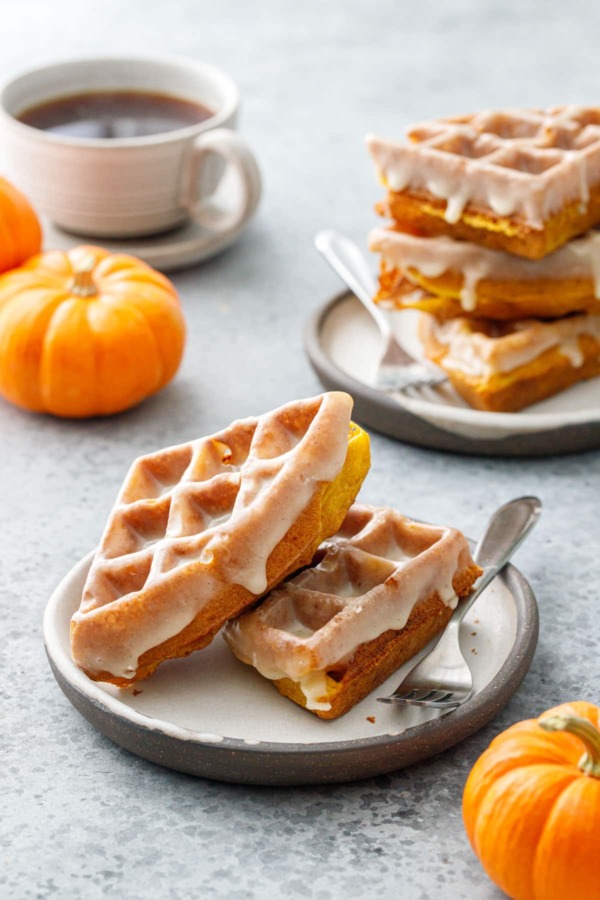 Two plates with stacks of Pumpkin Donut Waffles with a visibly drippy Vanilla Cardamom Glaze, mini pumpkins and cup of coffee in the background.