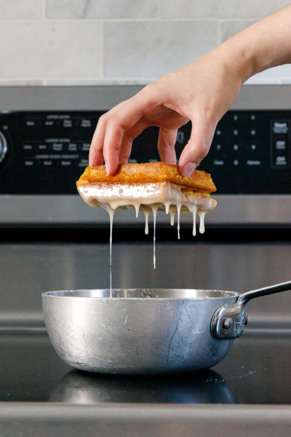 Dipping a pumpkin donut waffle into a small pan with vanilla caradmom glaze, letting the excess drip off.