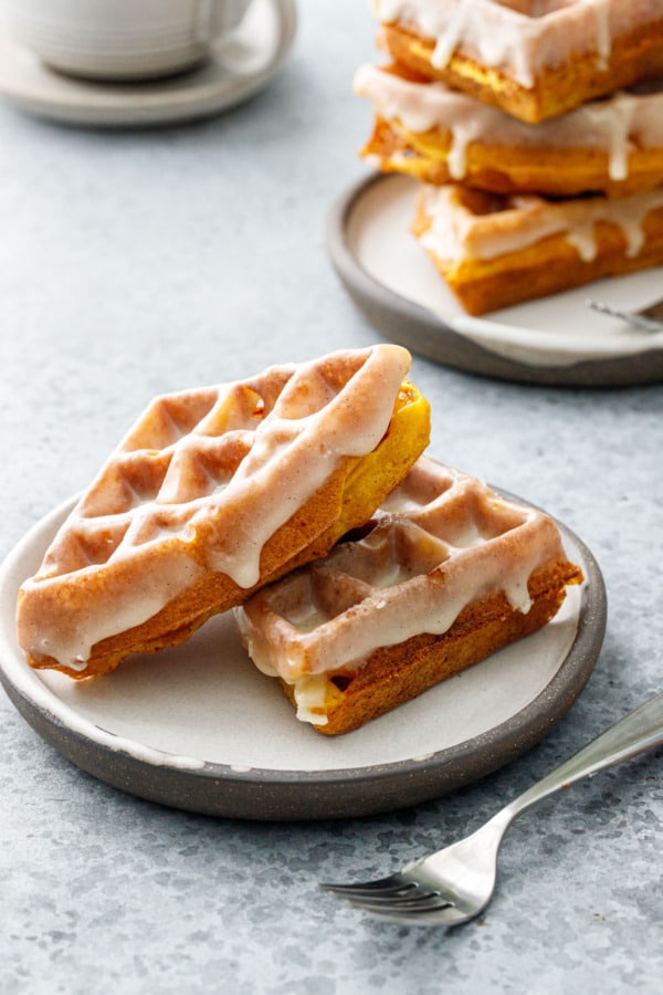 Two plates with stacks of Pumpkin Donut Waffles with Vanilla Cardamom Glaze, fork and coffee cup in the background.
