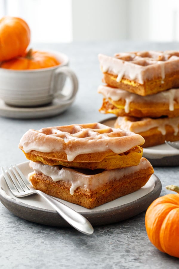 Two plates stacked with Pumpkin Donut Waffles with a visibly drippy Vanilla Cardamom Glaze, with mini pumpkins.