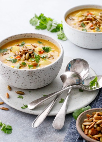 Two speckled ceramic bowls with Sausage & Corn Chowder, vintage spoons on the side with a bowl of toasted pepitas and loose cilantro leaves.