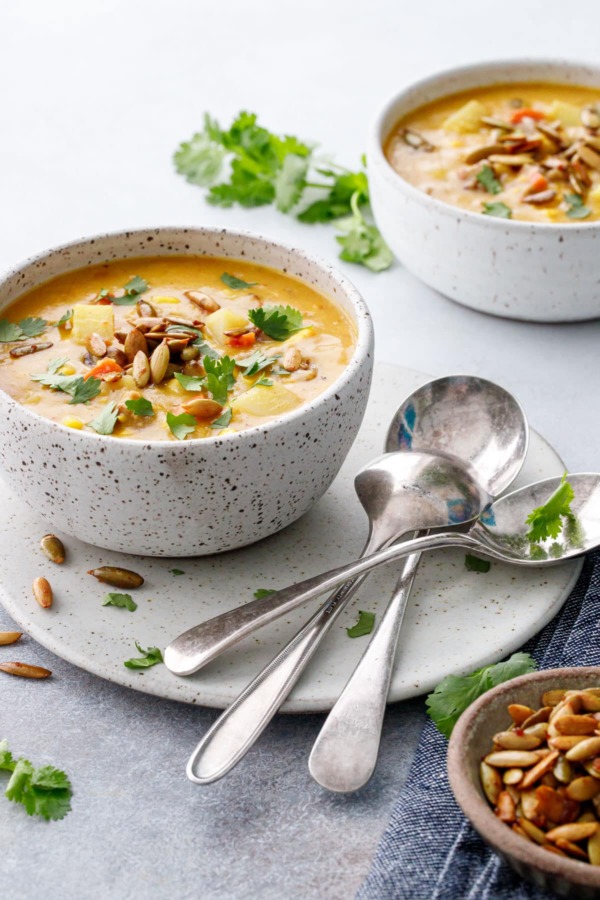 Two speckled ceramic bowls with Sausage & Corn Chowder, vintage spoons on the side with a bowl of toasted pepitas and loose cilantro leaves.