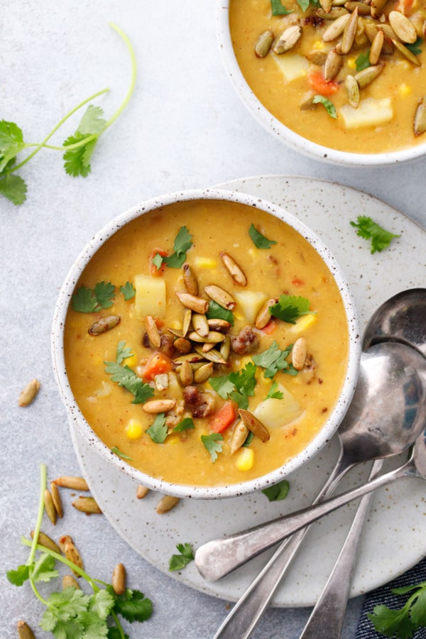 Overhead, two bowls of Sausage & Corn Chowder on a gray background, with silver spoons and scattered cilantro leaves.