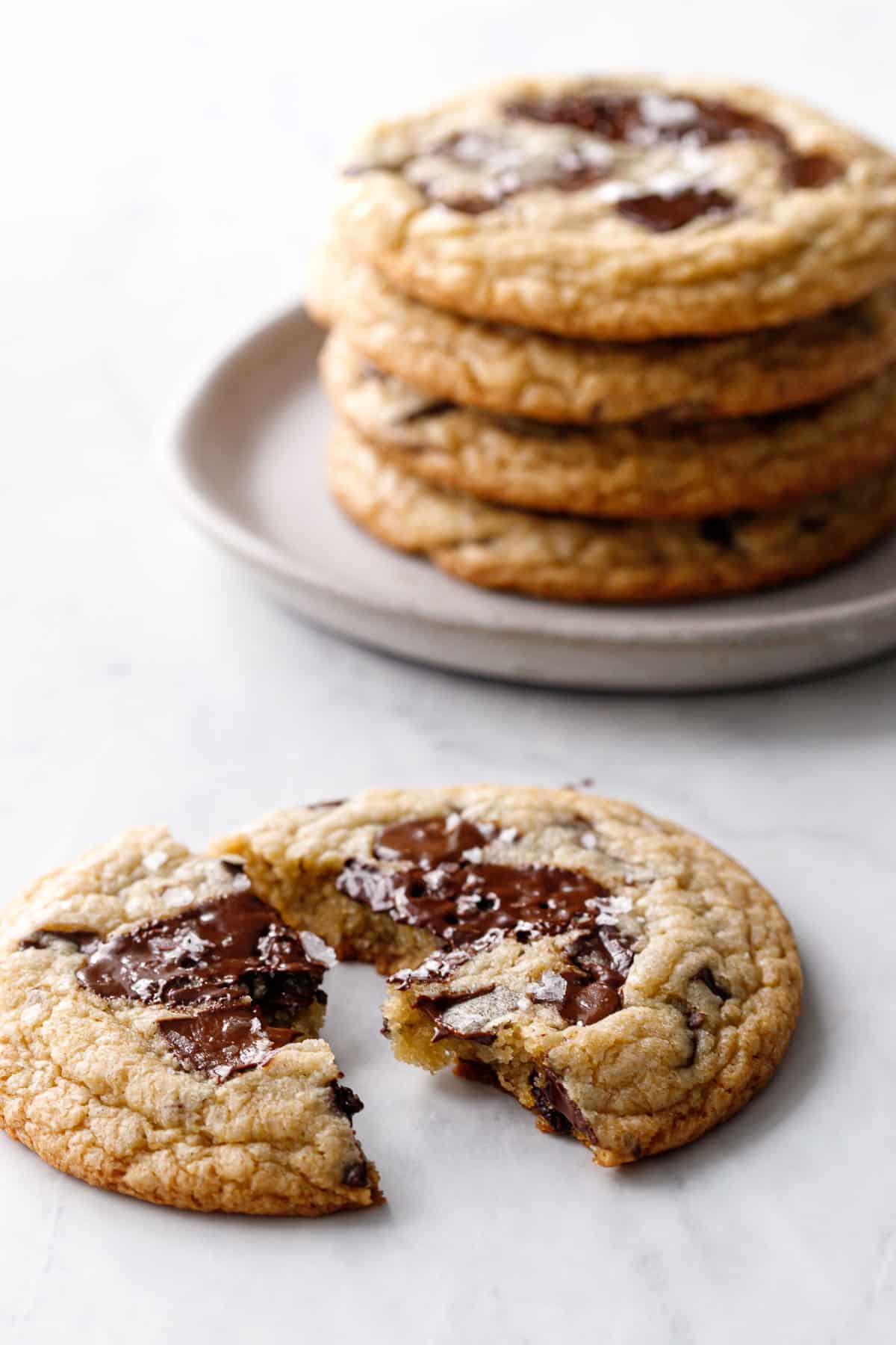 Amaretto Chocolate Chip Cookie broken in half to show gooey melty texture, with a stack of more cookies on a plate in the background.