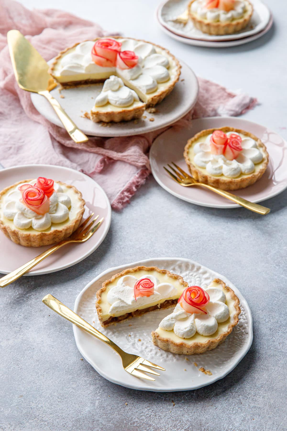 Messy scene featuring Caramel Apple Mousse Tarts, one big tart with a slice taken out and multiple smaller tartlets, one cut in half, with gold cutlery and a pink napkin.