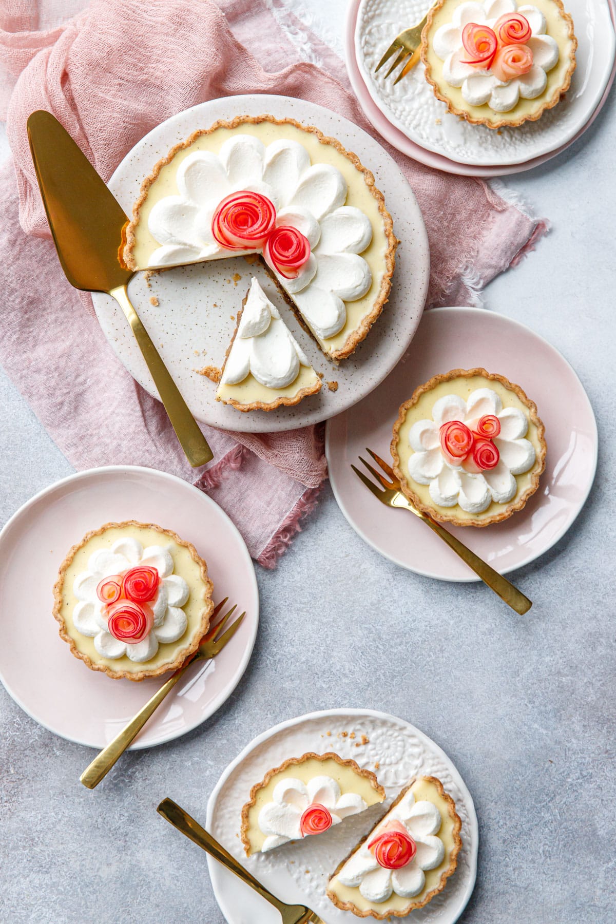 Overhead, messy scene featuring Caramel Apple Mousse Tarts, one big tart with a slice taken out and multiple smaller tartlets, with gold cutlery and a pink napkin.