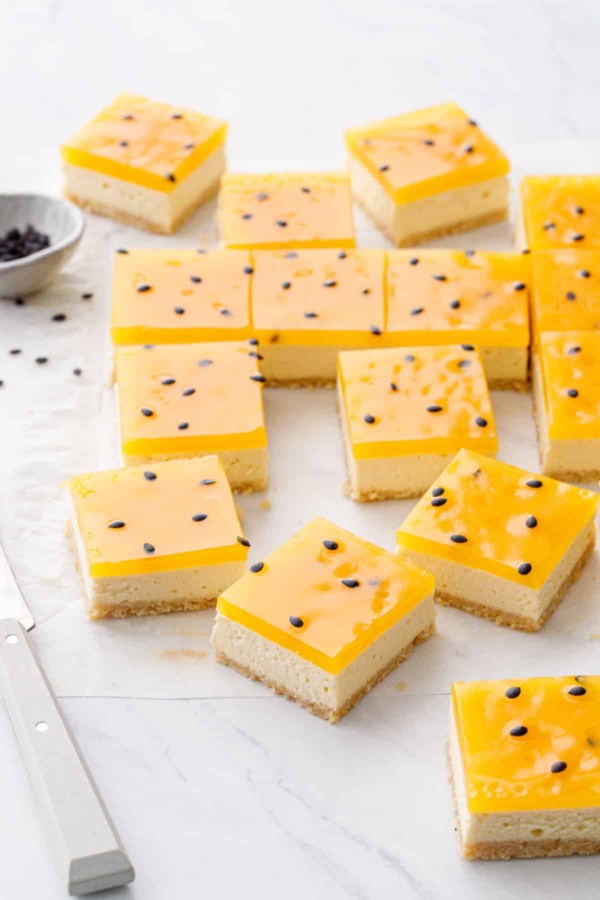 Square-cut Passionfruit Cheesecake Bars with a bright yellow top layer and black passionfruit seeds on a marble background.