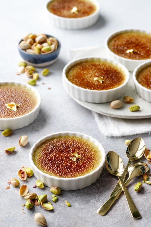 Six Pistachio Crème Brûlées in white ramekins, on gray background with gold spoons and scattered pistachios.