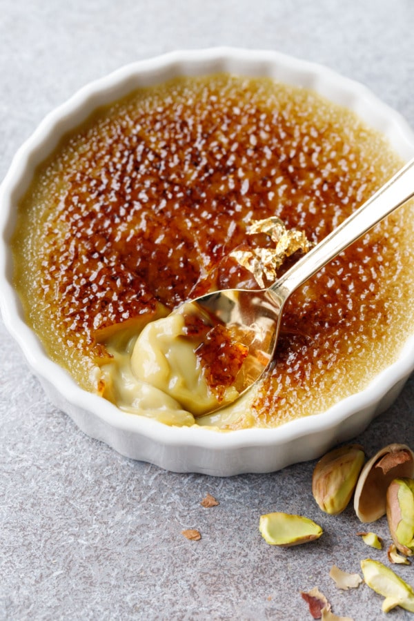 Closeup, gold spoon scooping a bite out of a ramekin of Pistachio Crème Brûlée, topped with a gold leaf garnish.