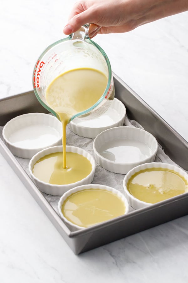 Pouring light green colored Pistachio Crème Brûlée into white fluted ramekins, in a baking dish.