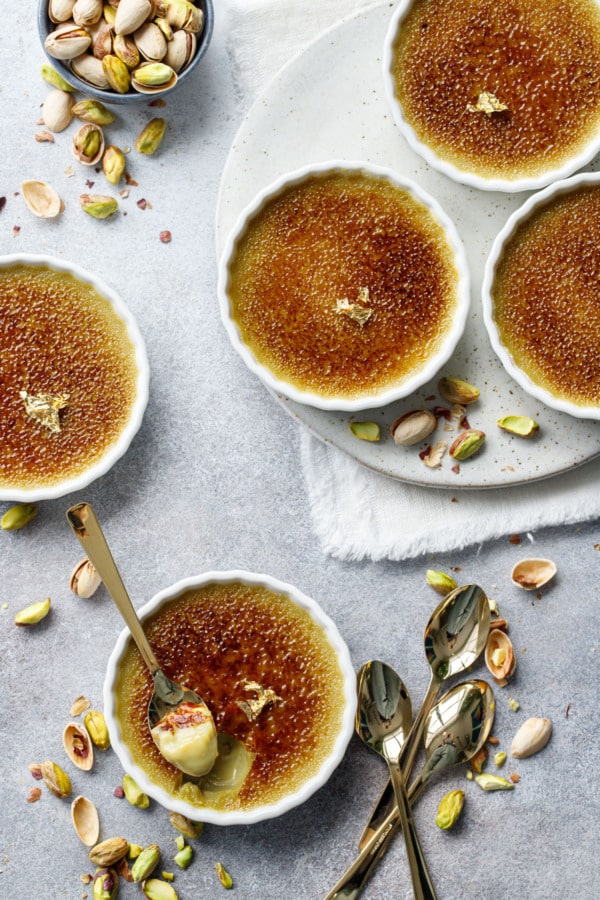Overhead, fluted white ramekins with Pistachio Crème Brûlée, one with a spoonful out of it to show texture, with gold leaf garnish on top.