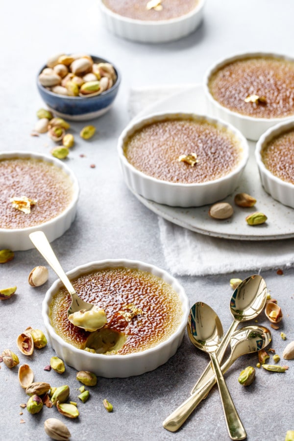 Scene with Pistachio Crème Brûlée in fluted ramekins, one with a spoonful out of it, messy pistachios scattered around and a few extra gold spoons.