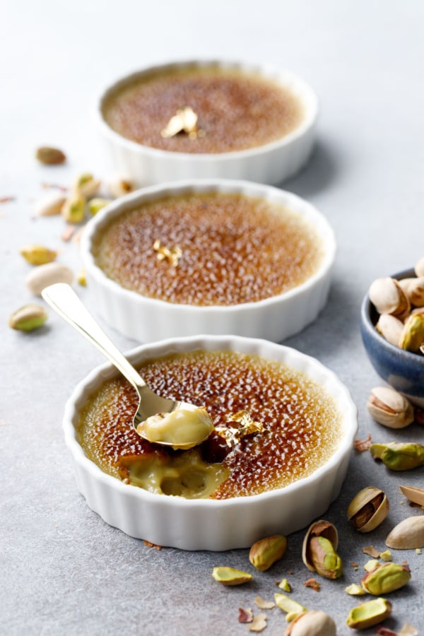 Three ramekins of Pistachio Crème Brûlée lined up, shallow depth of focus and raw pistachios scattered around.