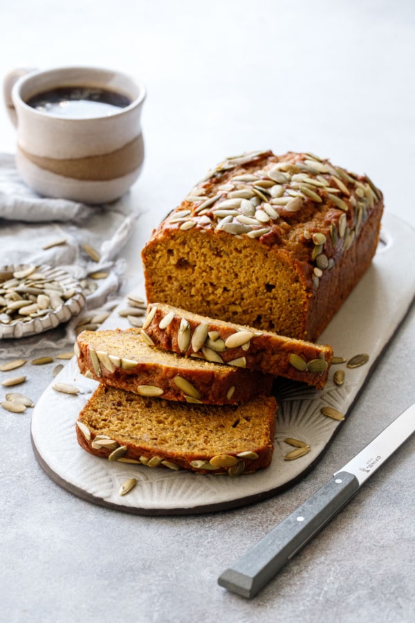 Sliced Spiced Pumpkin Banana Bread on a ceramic serving dish, with knife, cup of coffee and dish of pepitas on a gray background.