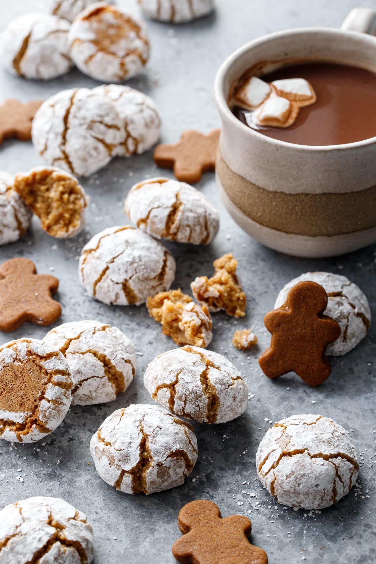 Messy arrangement of Gingerbread Amaretti Cookies and mini gingerbread men to visually indicate the flavor, plus a mug of hot chocolate in the background.