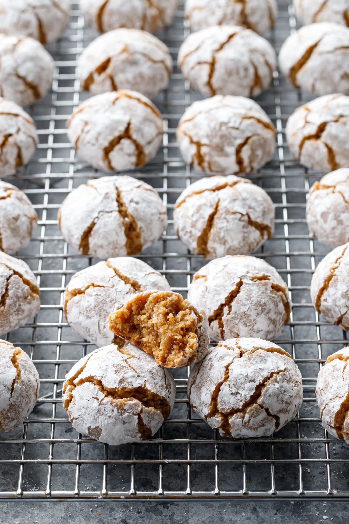 Wire rack with even rows of Gingerbread Amaretti Cookies, one split cookie sitting on top to show the interior texture.