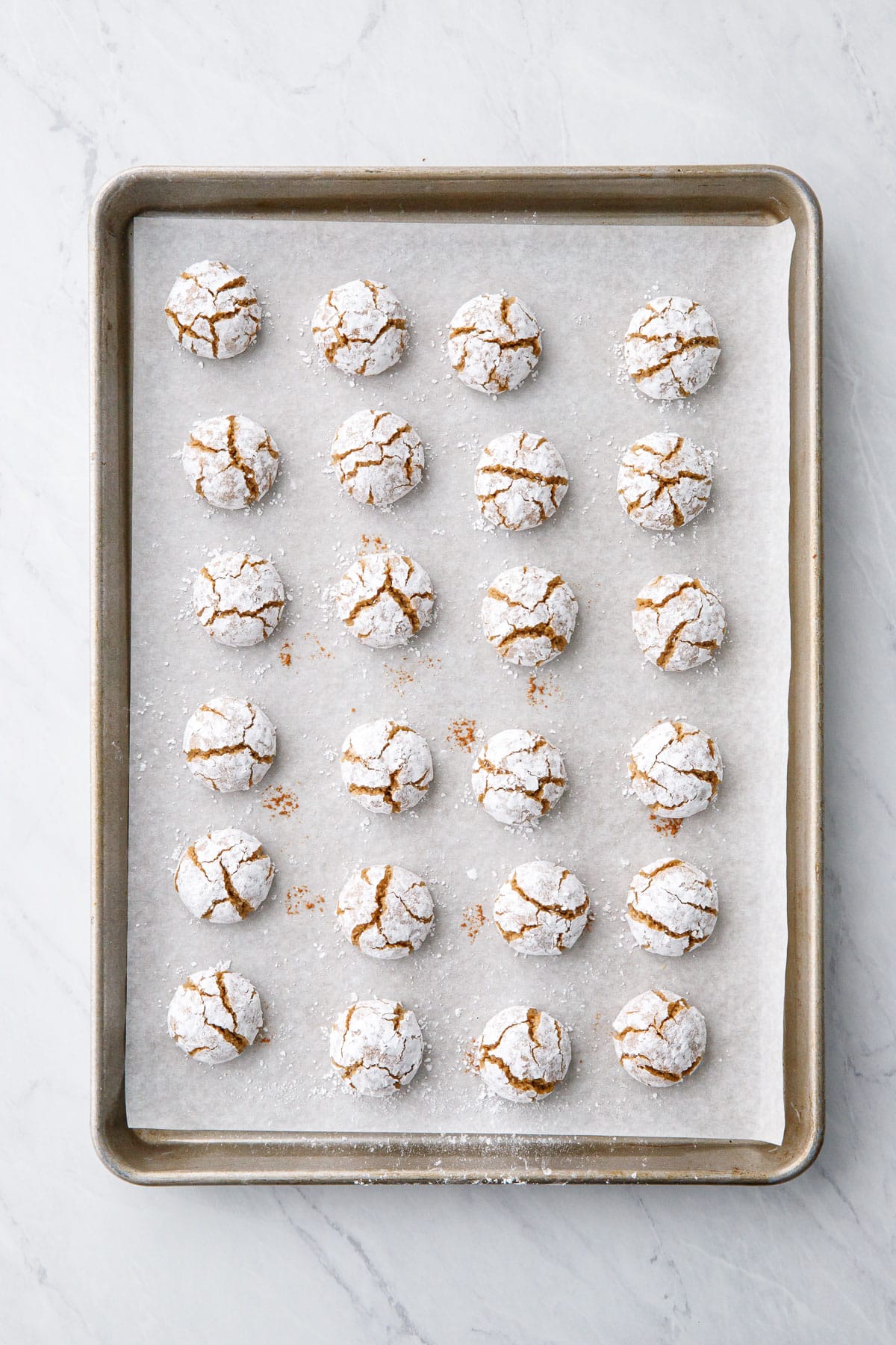 Overhead, parchment-lined baking sheet with rows of baked Gingerbread Amaretti Cookies; the cookies are coated in powdered sugar with a crinkled appearance.