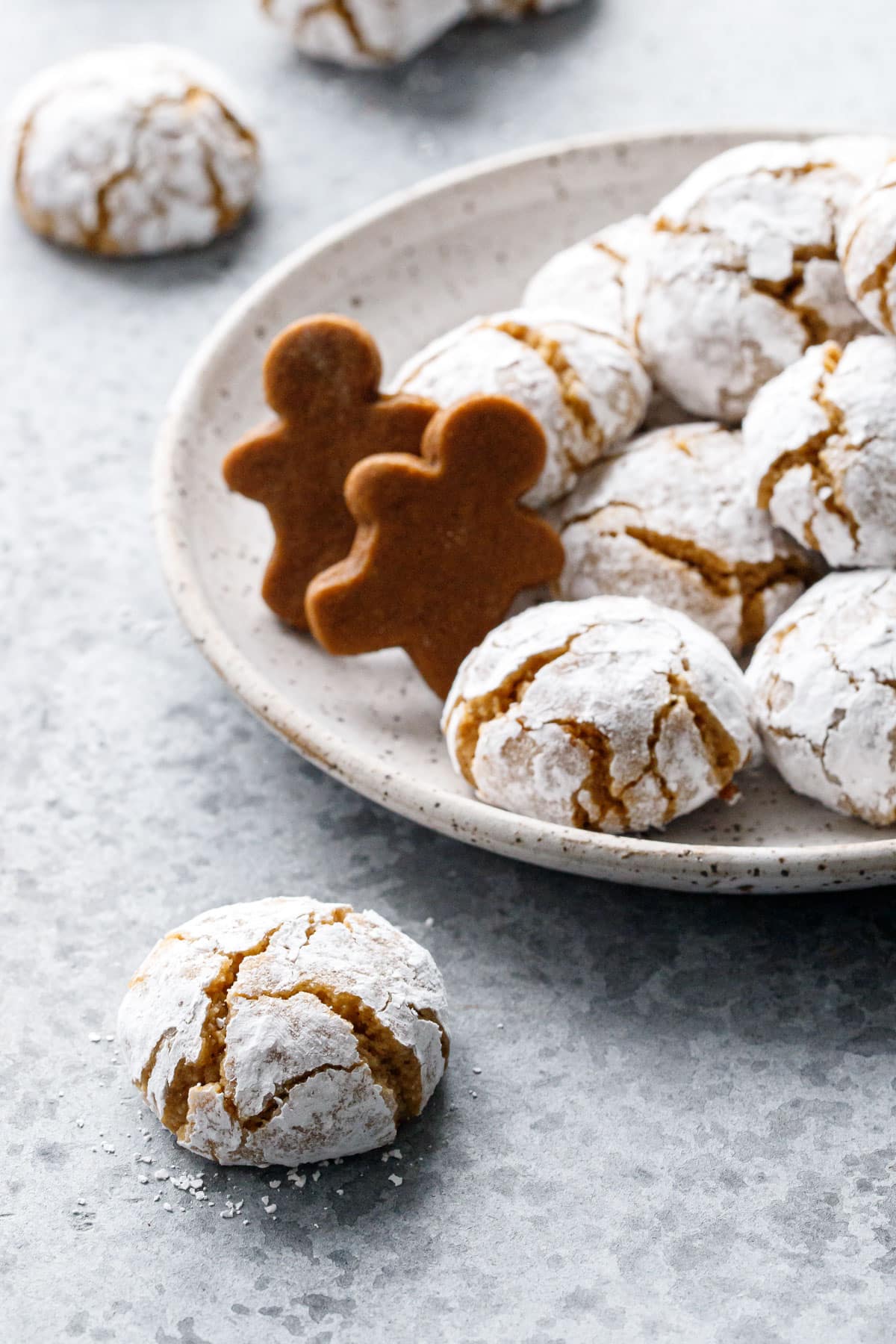 Closeup showing the crinkle appearance and powdered sugar coating of a Gingerbread Amaretti Cookie in the foreground, plate with more cookies and two small gingerbread men in the background.