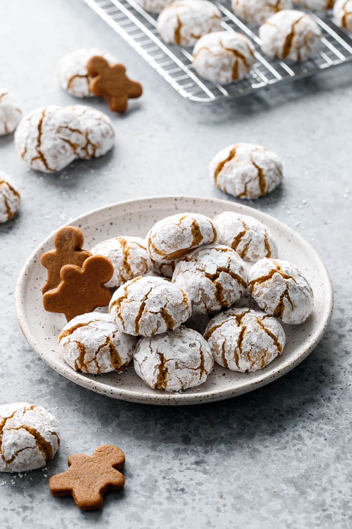 Plate stacked with a pile of Gingerbread Amaretti Cookies, plus a few small gingerbread men and more cookies scattered around.