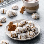 Plate with pile of Gingerbread Amaretti Cookies, more cookies scattered around plus a mug of hot chocolate.