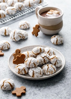 Plate with pile of Gingerbread Amaretti Cookies, more cookies scattered around plus a mug of hot chocolate.