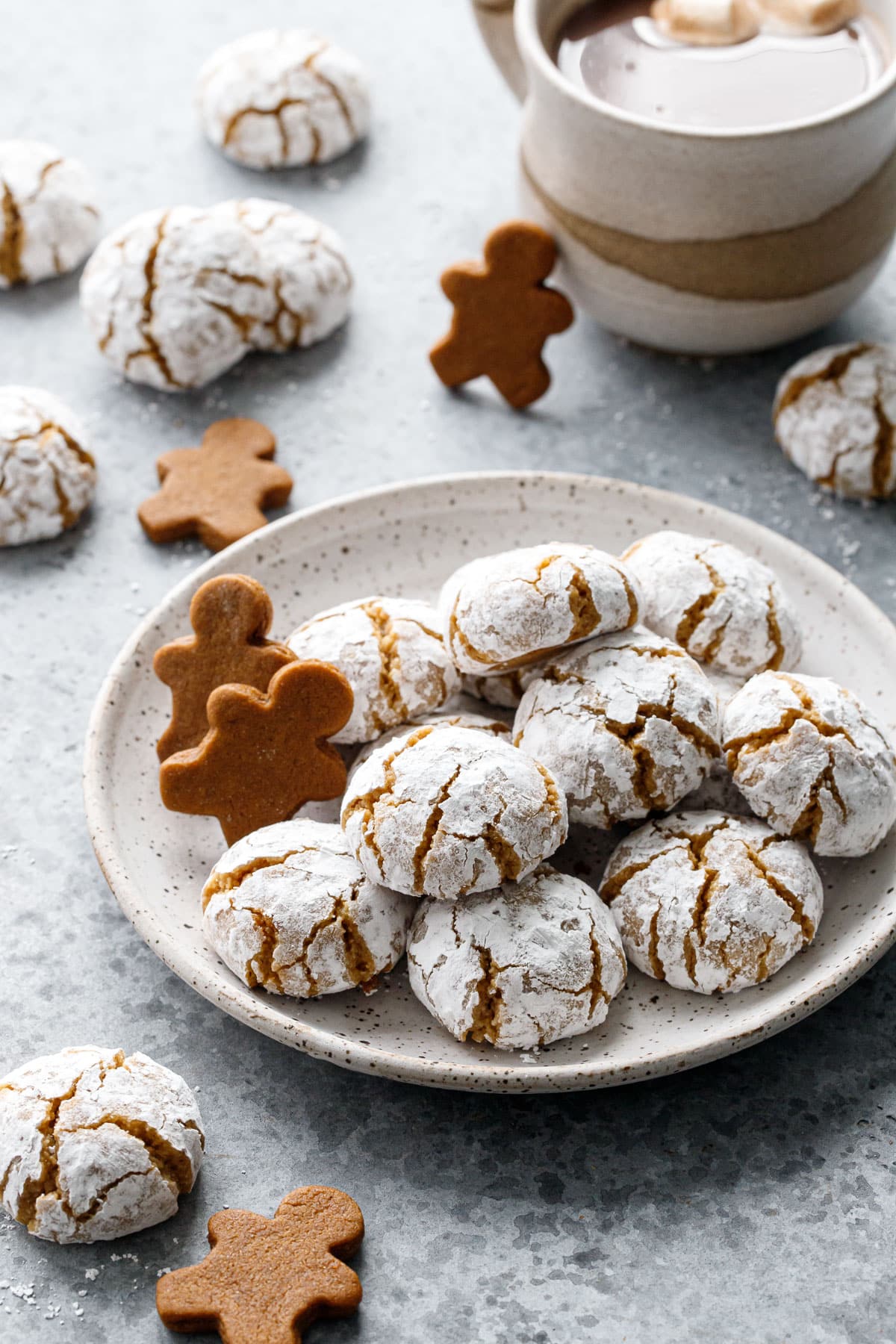 Plate with pile of Gingerbread Amaretti Cookies, with more cookies, a mug of hot chocolate, and mini gingerbread men scattered around.