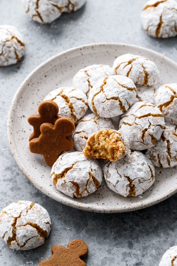 Plate with pile of Gingerbread Amaretti Cookies, one split open to show the soft and chewy texture within, plus gingerbread men to visually indicate the cookies' flavor.