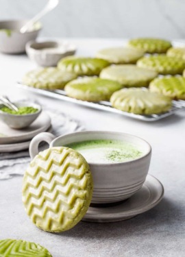 Cup of matcha with a zig-zag stamped matcha cookie leaning up against it, small bowls and a cooling rack with more cookies in the background.