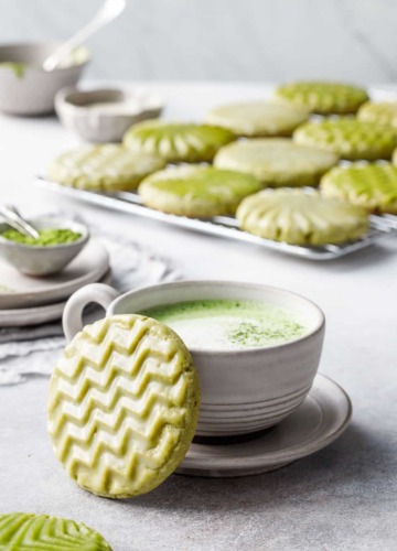 Cup of matcha with a zig-zag stamped matcha cookie leaning up against it, small bowls and a cooling rack with more cookies in the background.