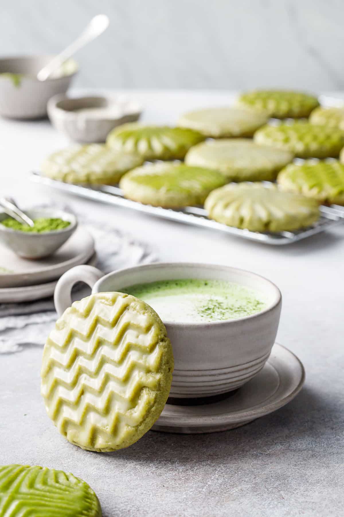 Cup of matcha with a zig-zag stamped matcha cookie leaning up against it, small bowls and a cooling rack with more cookies in the background.