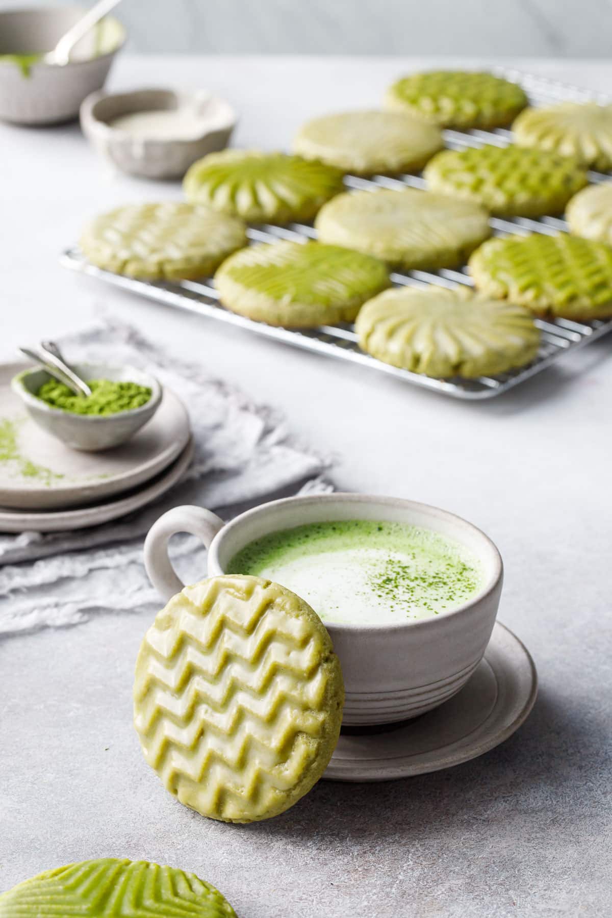 Cup of matcha with a zig-zag stamped matcha cookie leaning up against it, small bowls and a cooling rack with more cookies in the background.