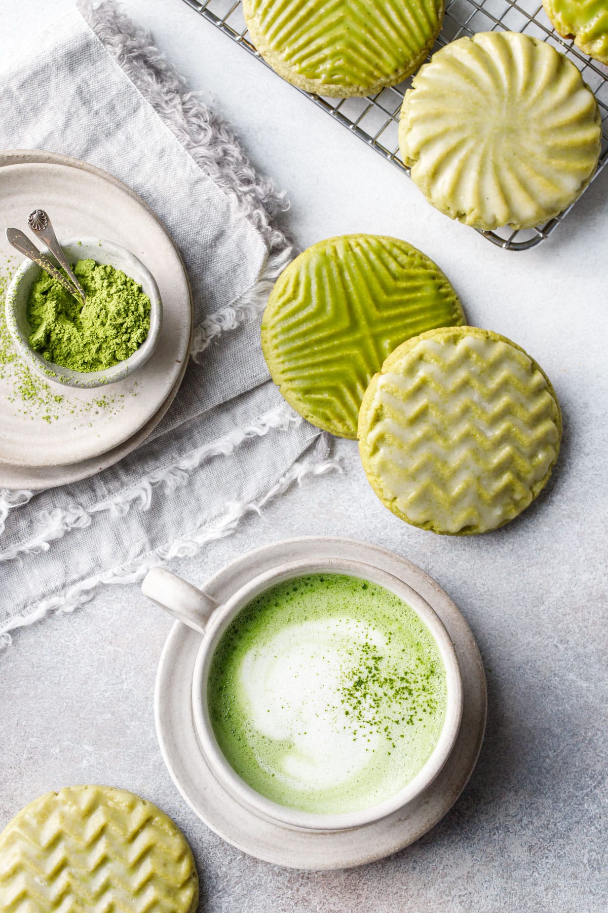Overhead, geometric stamped matcha sugar cookies with green and white glaze, mug of foamy matcha latte and a small bowl of matcha powder all on a gray background.