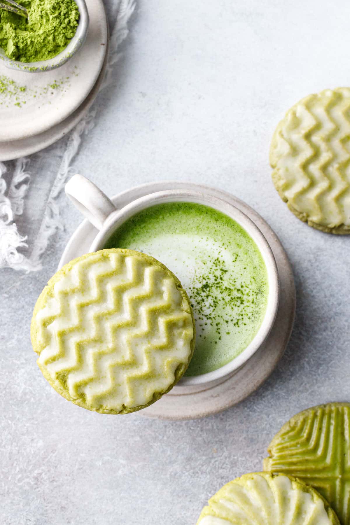 Overhead, glazed matcha sugar cookie with a zig zag design resting on the rim of a mug of matcha latte, more cookies and a small bowl of matcha powder in the background.