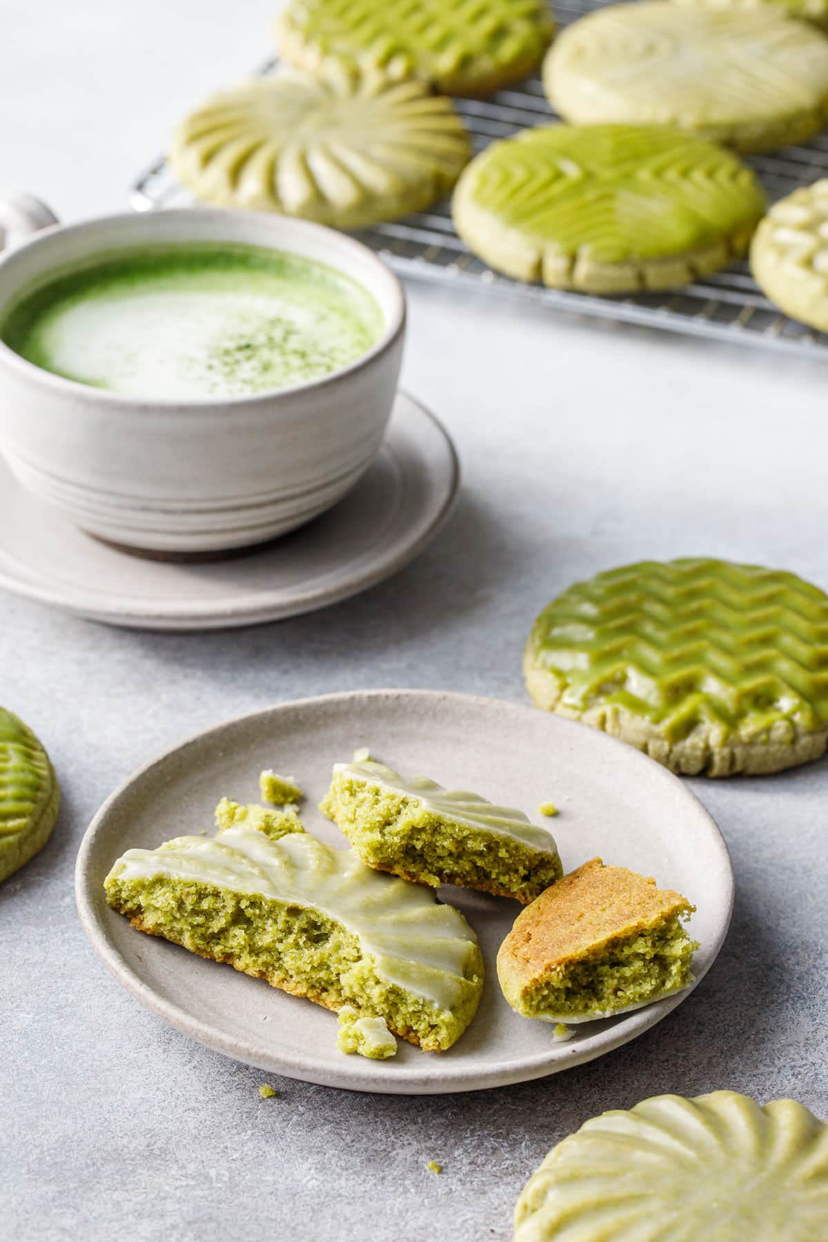 Ceramic plate with a Glazed Matcha Sugar Cookie broken in pieces to show the inner texture, mug of matcha and more cookies in the background.