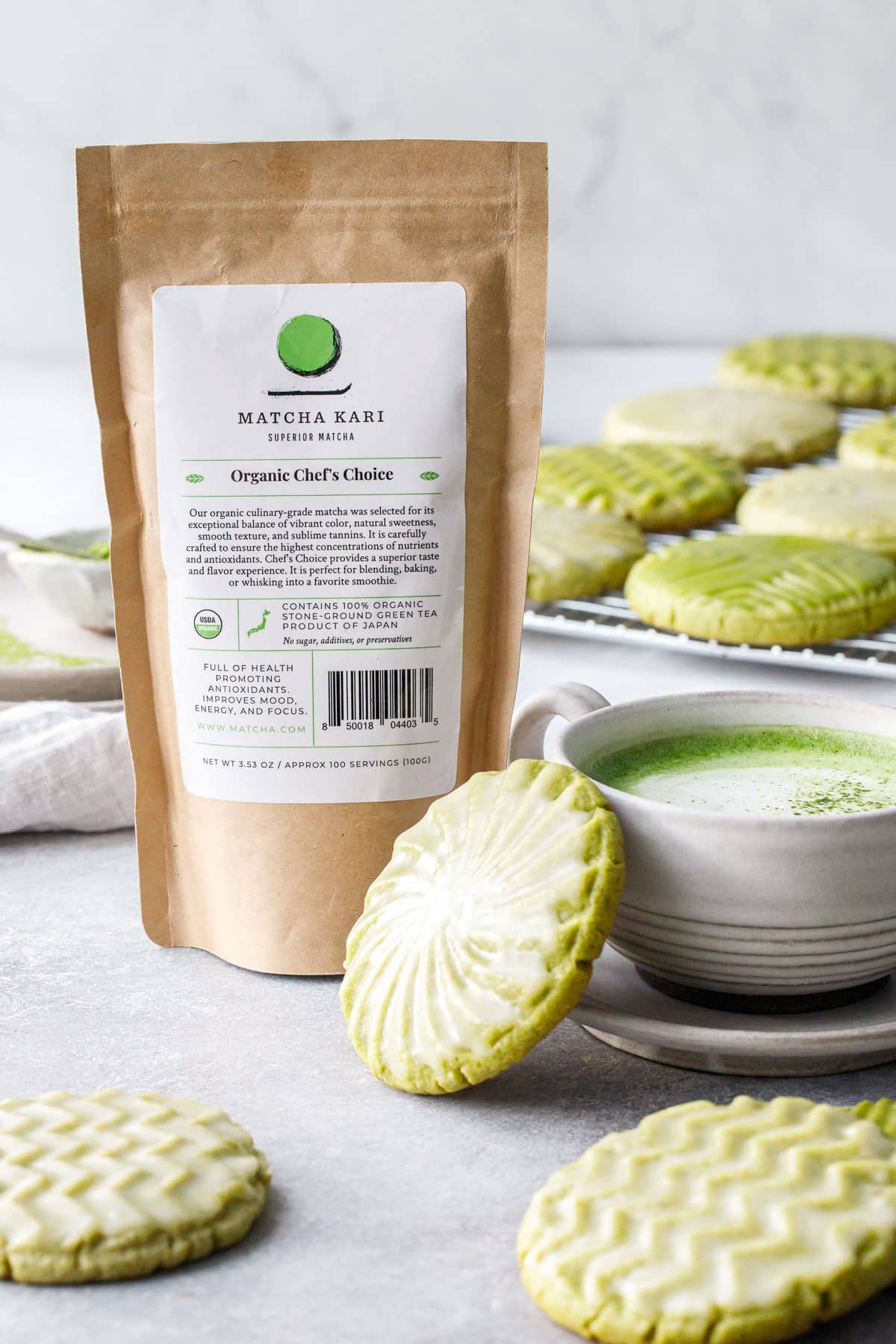 Glazed Matcha Sugar Cookies and a brown bag of Matcha Kari culinary matcha used to make the cookies, plus a mug of matcha latte and more cookies in the background.