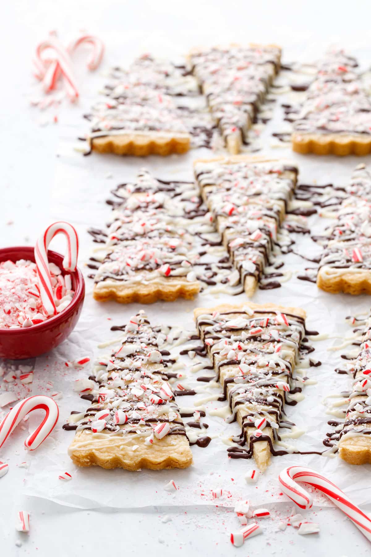 Rows of triangular Peppermint Bark Shortbread with white and dark chocolate drizzle and topped with crushed candy canes, and a red bowl of candy cane pieces and a few mini candy canes on the side.