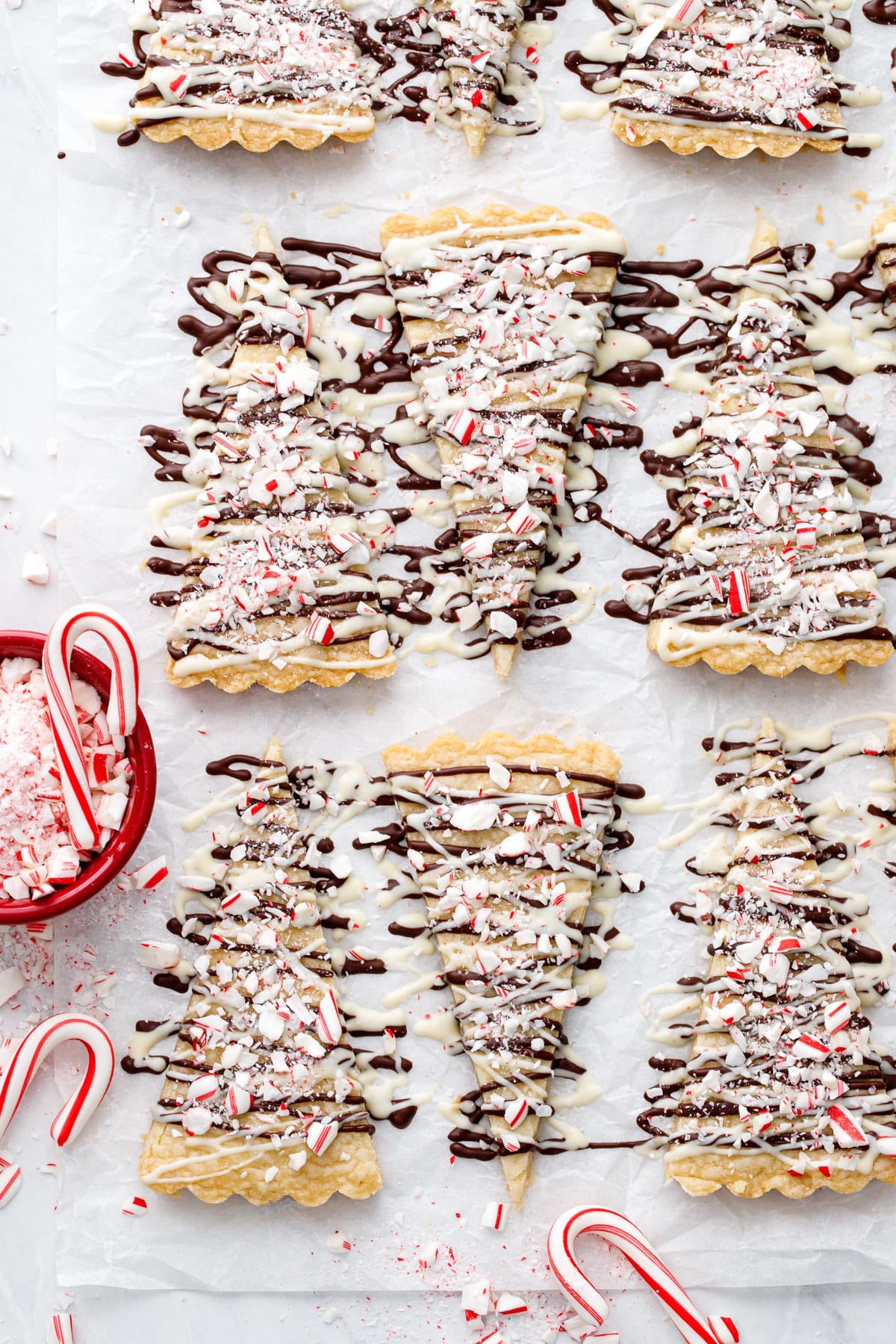 Overhead, rows of triangular shaped Peppermint Bark Shortbread on white parchment, with a bowl of crushed candy canes and whole mini candy canes on the side.
