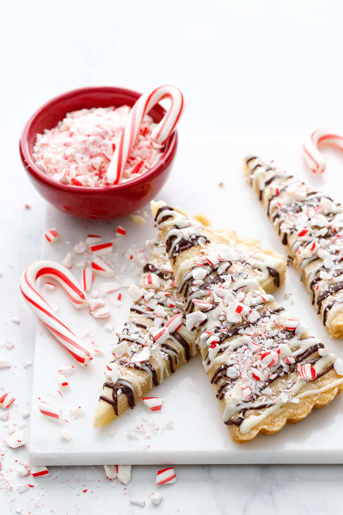 Peppermint Bark Shortbread cookies cut into triangle/christmas-tree shapes, messy arrangement on a marble square with crushed and whole candy canes scattered around.