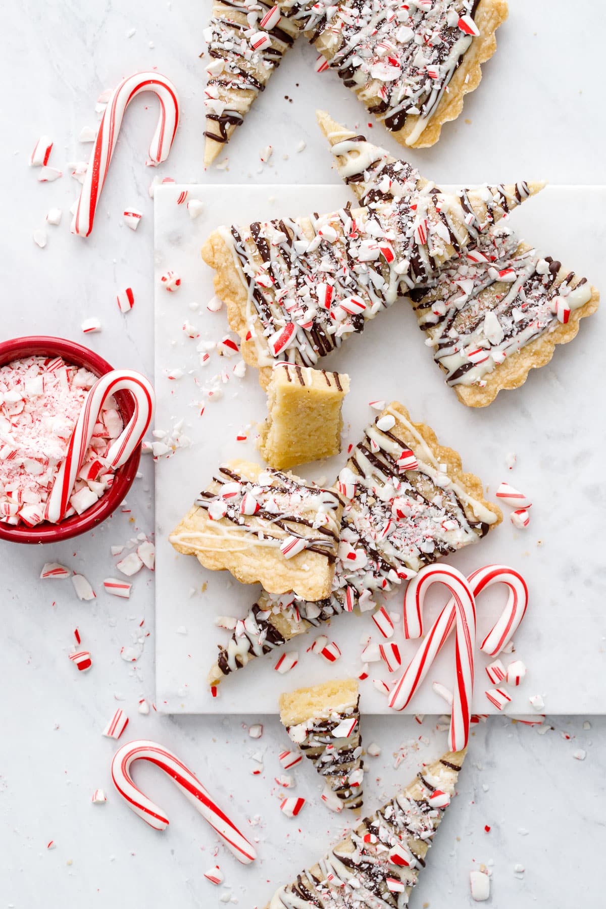 Overhead, messy arrangement of Peppermint Bark Shortbread cookies on a marble background, with crushed and whole mini candy canes scattered around.