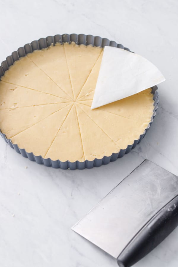 Tart pan with shortbread dough, divided into 12 even wedges, showing the parchment triangle used as a template and the bench scraper used for scoring.