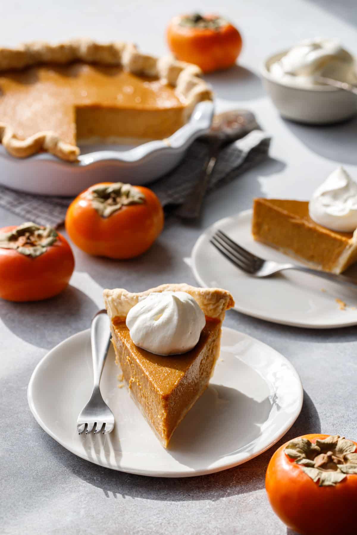 Slices of Persimmon Pie on white plates, what's left of the full pie in the background along with a few Fuyu persimmons.