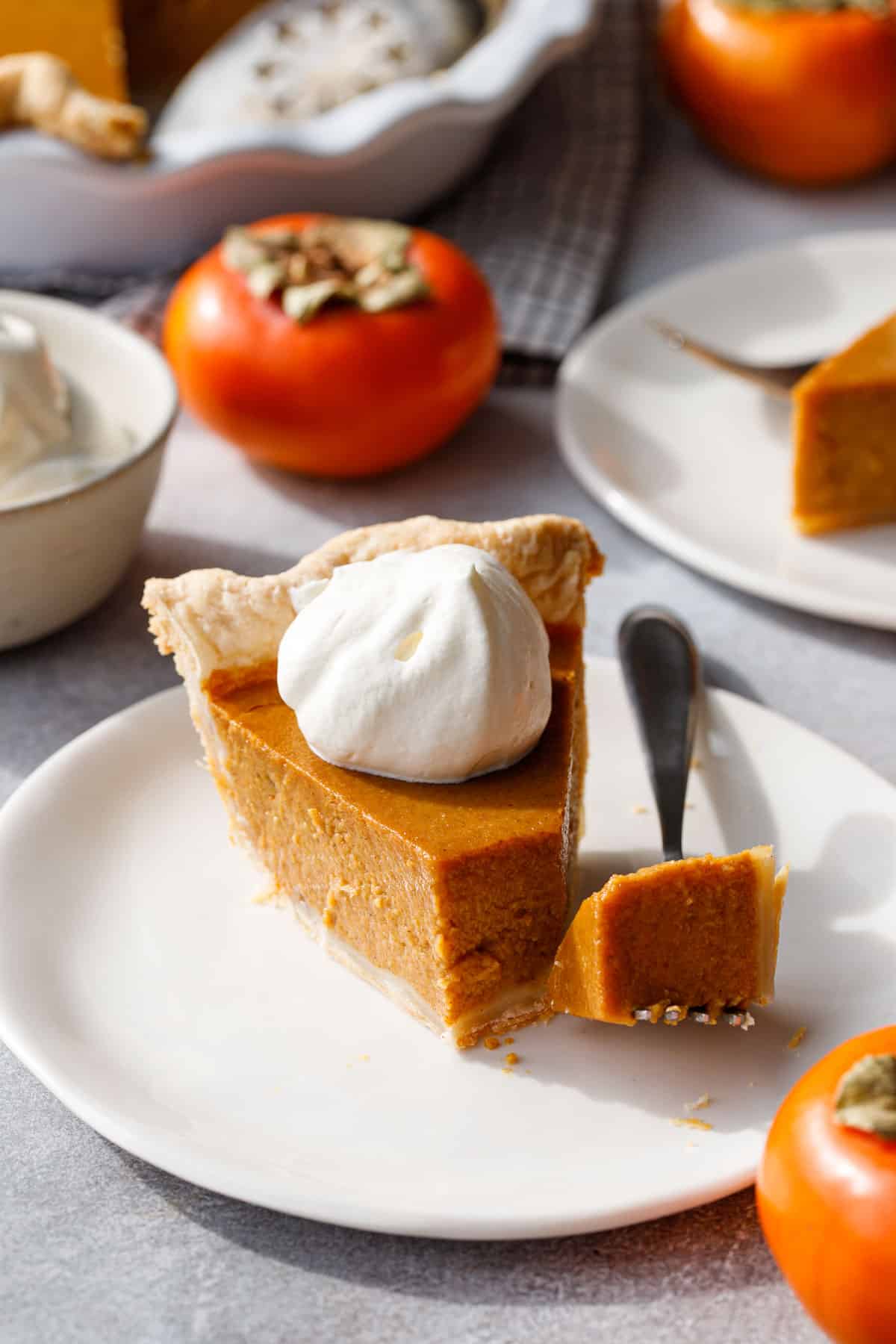 Bite resting on a fork with a slice of Persimmon Pie on a white plate, orange persimmons and a bowl of whipped cream.