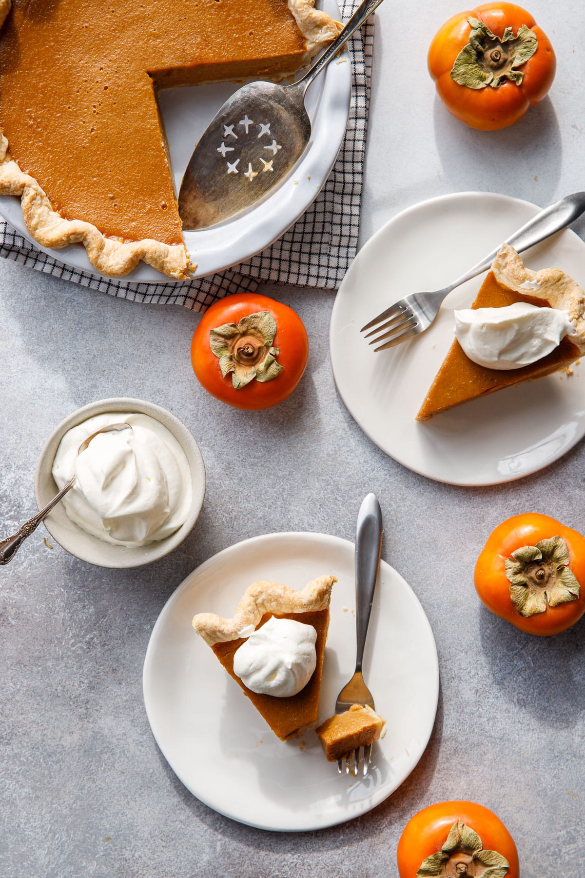 Overhead, two slices of Persimmon Pie on plates plus the rest of the whole pie, with Fuyu persimmons and a bowl of whipped cream.