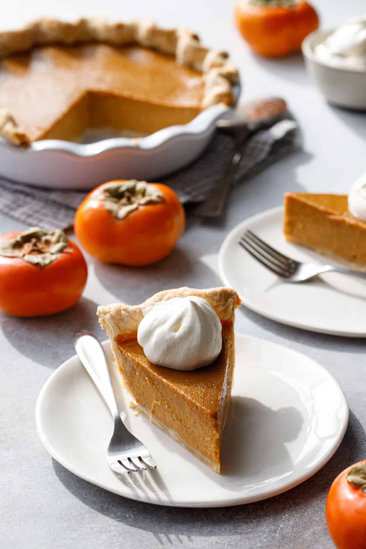 Slice of Persimmon Pie on a white plate with fork and perfect dollop of whipped cream on top; whole persimmons and the rest of the pie in the background.
