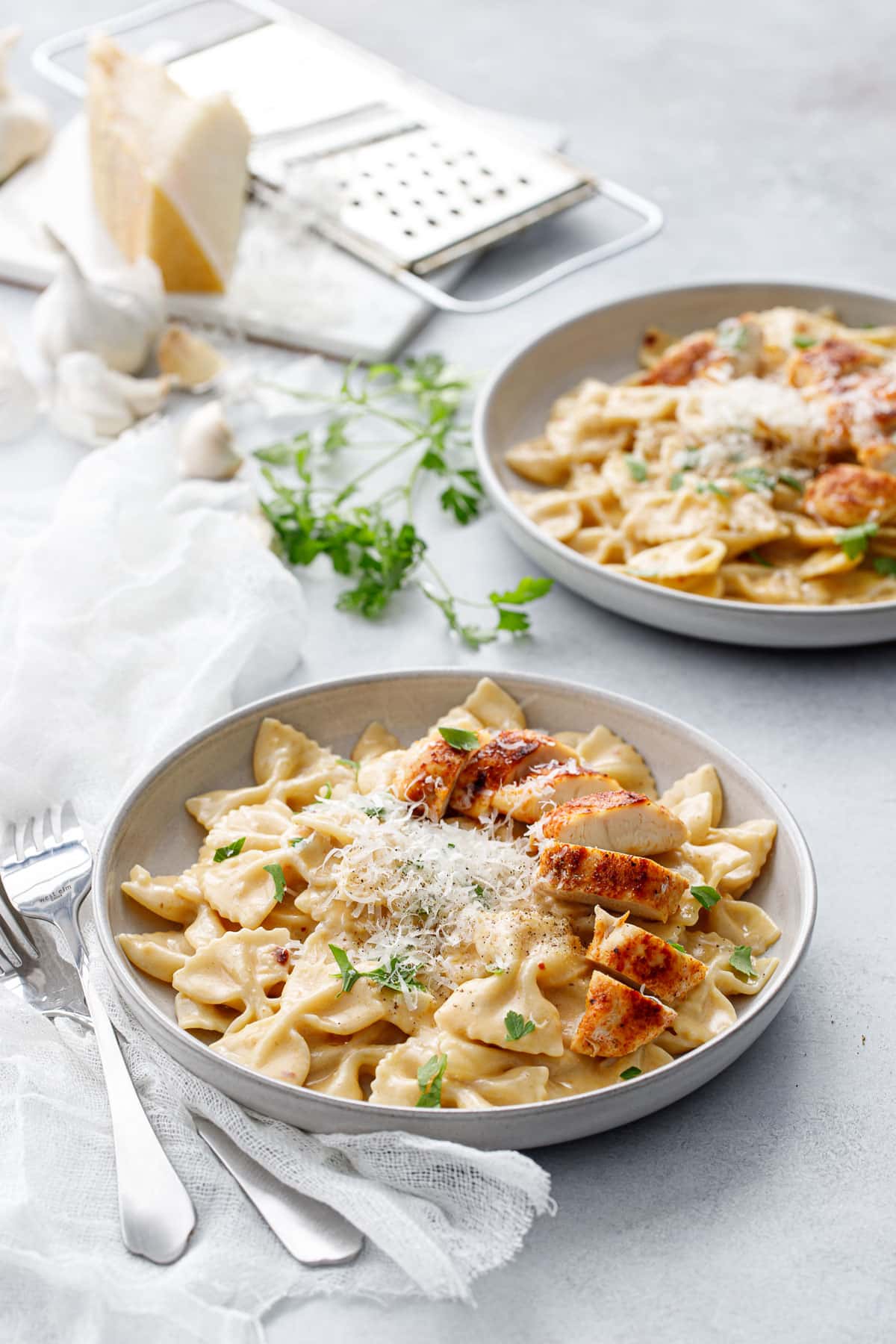 Two shallow bowls with Creamy Garlic Chicken Pasta, napkin and forks and a piece of parmesan cheese in the background.