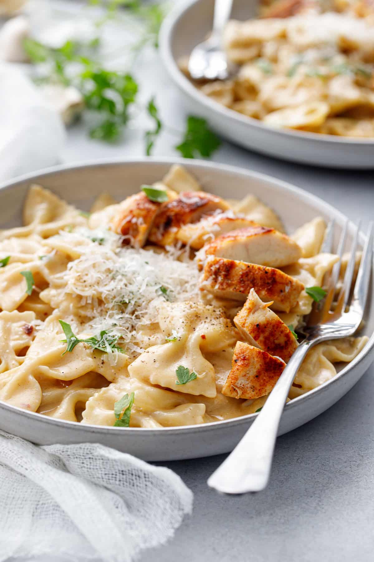 Closeup of bowl of creamy farfalle pasta topped with browned chicken tenders cut into pieces, with little pieces of parsley and a pile of grated parmesan cheese.