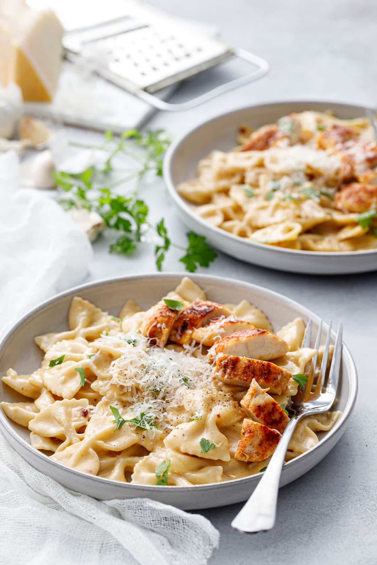 Two shallow bowls with Creamy Garlic Chicken Pasta, napkin and forks and a piece of parmesan cheese in the background.