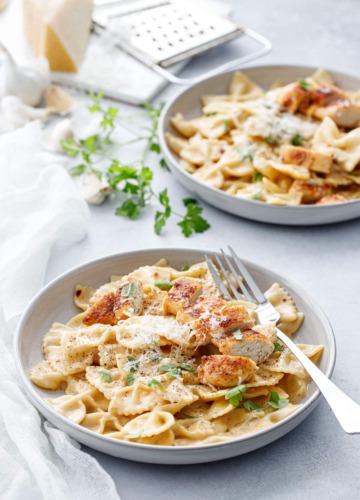 Two shallow bowls with Creamy Garlic Chicken Pasta, napkin and forks and a piece of parmesan cheese in the background.