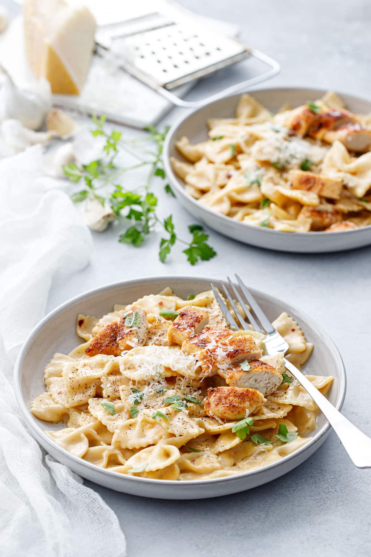 Two shallow bowls with Creamy Garlic Chicken Pasta, napkin and forks and a piece of parmesan cheese in the background.