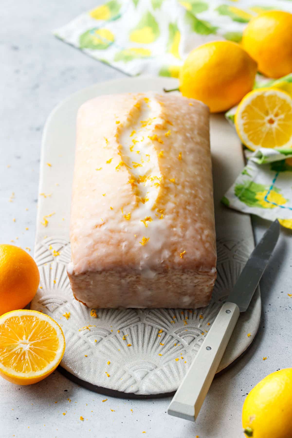 Meyer Lemon Olive Oil Loaf Cake sitting on a textured ceramic plate, with lemon print napkin and whole/halved meyer lemons scattered around.